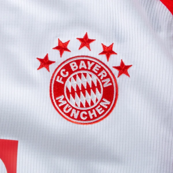 A close-up of the FC Bayern München logo on the white 23/24 Long Sleeve Home Jersey. The circular emblem includes red text and elements, topped by four red stars.