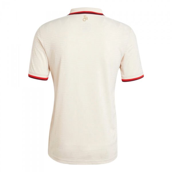 The image displays the rear of a cream-colored, short-sleeve Bayern Munich Third Jersey 24/25, featuring red sleeve and collar accents and a small embroidered logo near the back collar.