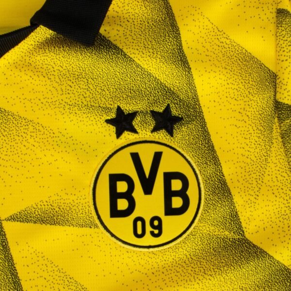 Close-up of the Borussia Dortmund Third Jersey 23/24 features a yellow and black design with two black stars on the circular BVB 09 emblem, against a background highlighted by a geometric pattern with textured black dots.
