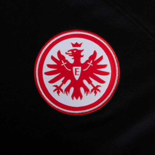 The Eintracht Frankfurt Away Jersey 23/24 features a symmetrical and stylized red eagle with a crown and the letter E in the center, set within a red circle on a black background, emphasizing its wings and talons.