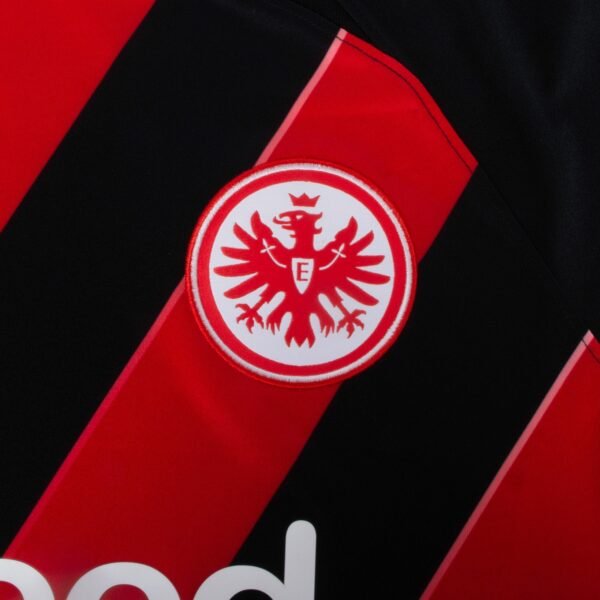 Close-up of the Eintracht Frankfurt Home Jersey 23/24, featuring red and black stripes and an embroidered emblem. The emblem displays a crowned eagle with outspread wings and the letter E on its chest.