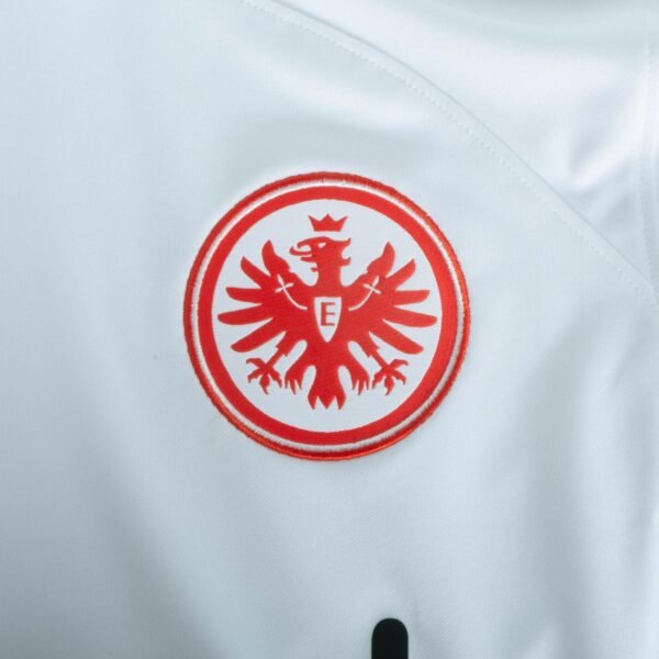 The Eintracht Frankfurt Third Jersey 23/24 features a white fabric with a bold red circular emblem. It showcases an eagle spreading its wings, crowned and adorned with an E, standing out against the plain background to highlight its intricate design and vibrant colors.