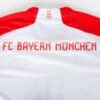 The FC Bayern Munich Home Jersey 23/24 boasts MIA SAN MIA elegantly near the neckline, with the team name prominently displayed in bold red letters across the back.