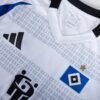 Close-up of the Hamburger SV Home Jersey 24/25, featuring the Adidas logo, a white fabric, the Hamburger SV badge with a star above it, and a partial geometric pattern in the background.