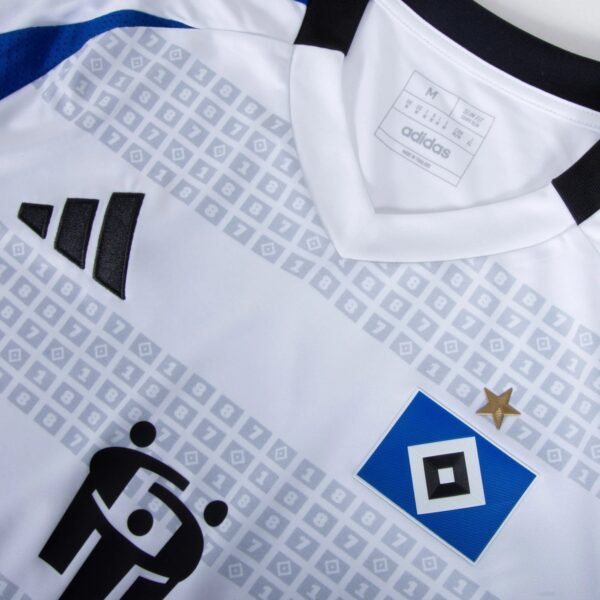 Close-up of the Hamburger SV Home Jersey 24/25, featuring the Adidas logo, a white fabric, the Hamburger SV badge with a star above it, and a partial geometric pattern in the background.